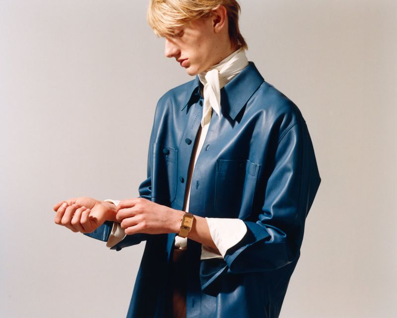 Young man in a blue leather jacket fastens cuff, posing indoors against light grey backdrop, showcasing fashion and a wristwatch.