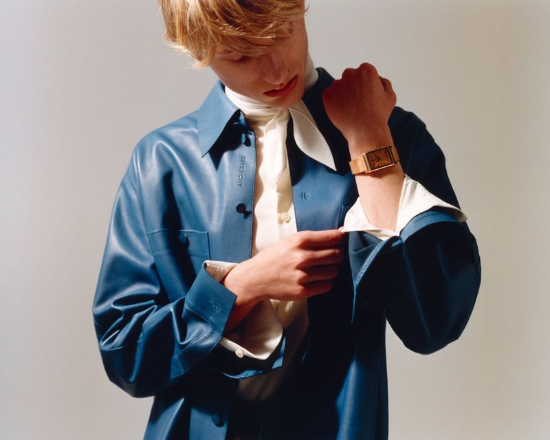Model wearing a blue leather shirt and gold watch adjusting cuffs against a seamless grey studio background