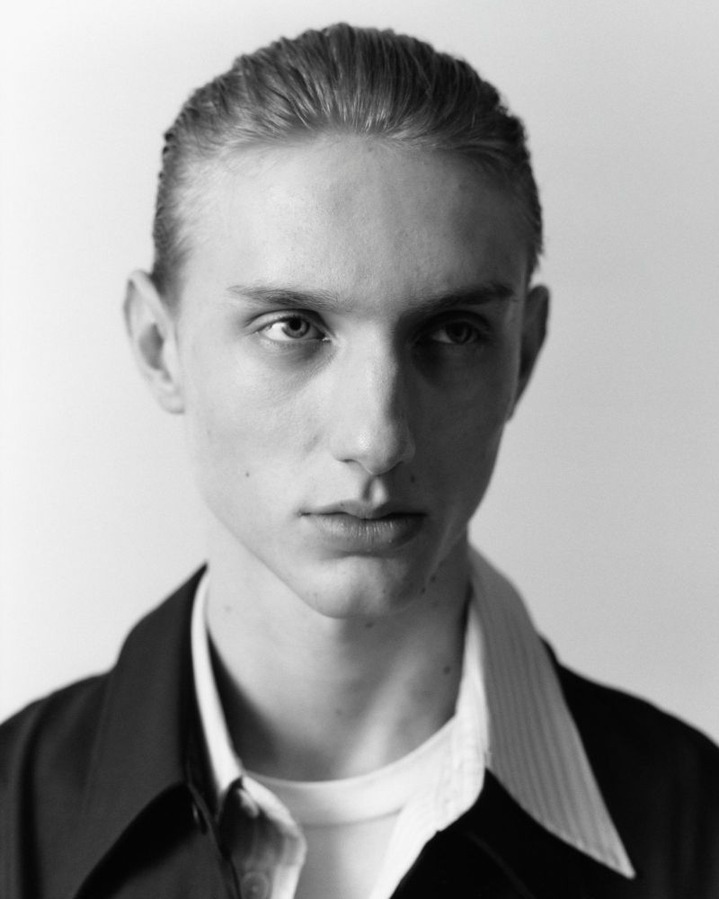 Close-up portrait of a young male model with slicked-back hair wearing a dark jacket and white shirt against a plain background.