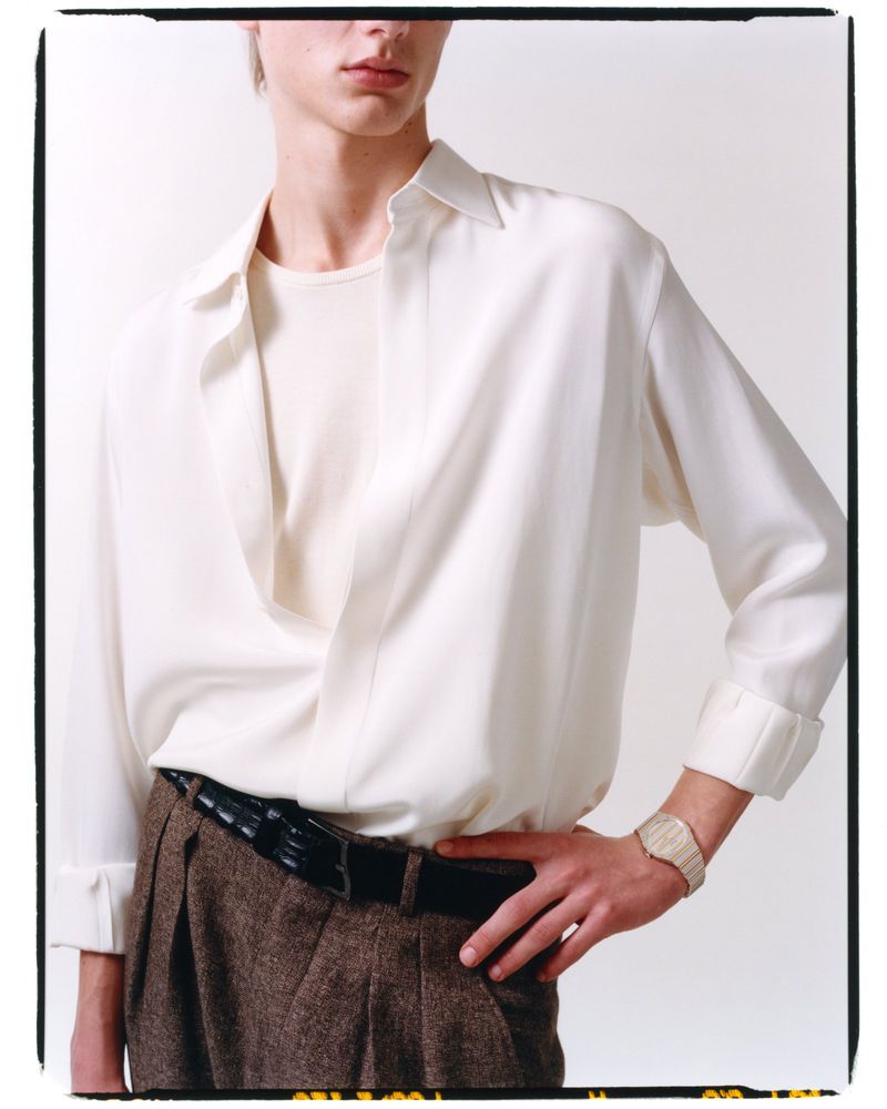 Model wearing a white shirt and brown trousers with a watch against a white background
