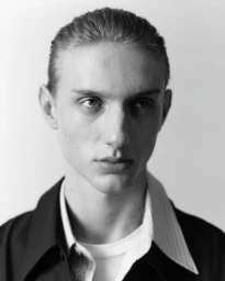 Close-up portrait of a young male model with slicked-back hair wearing a dark jacket and white shirt against a plain background.