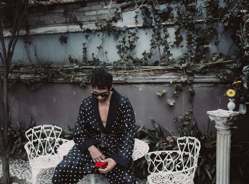 Actor Diego Calva wearing a black and white polka dot robe holding a red flower in an outdoor garden setting.