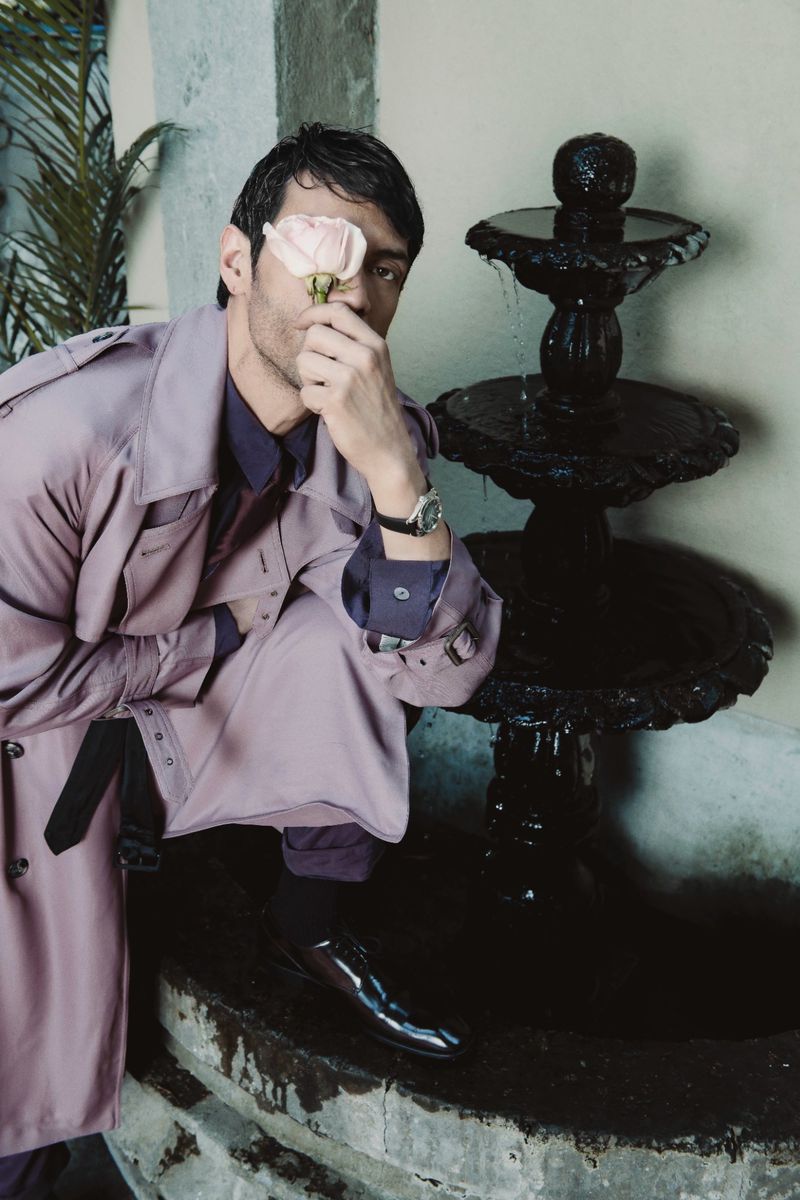 Man crouching by a stone fountain holding a pink rose to his eye wearing a lavender trench coat and dark trousers.
