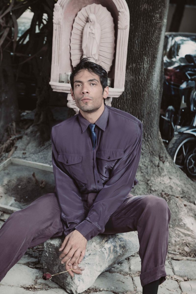 Actor Diego Calva seated outdoors wearing a purple shirt and trousers with a shrine and tree trunk in the background