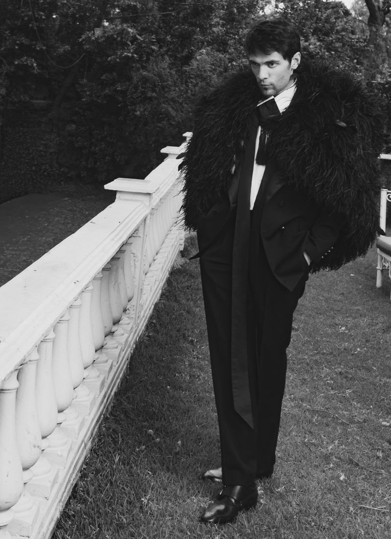 Man wearing a black feathered coat and tuxedo standing outdoors next to a white stone balustrade