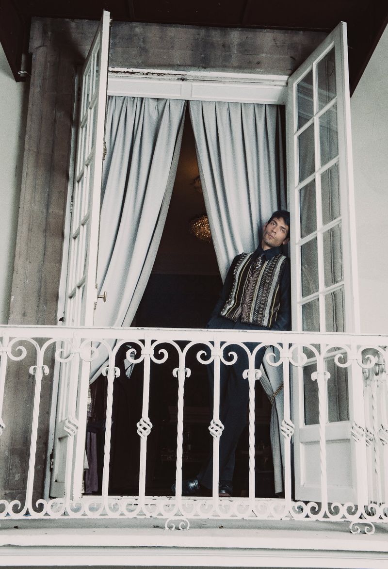 Diego Calva leaning in a balcony doorway wearing a patterned vest and dark trousers for Man About Town.