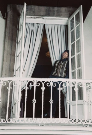 Diego Calva leaning in a balcony doorway wearing a patterned vest and dark trousers for Man About Town.