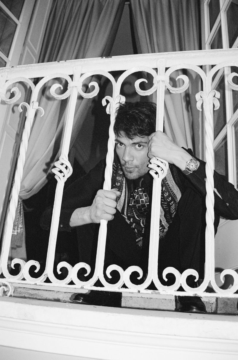 Black and white portrait of a man behind a white decorative iron railing, looking at the camera with a patterned scarf and dark clothing.