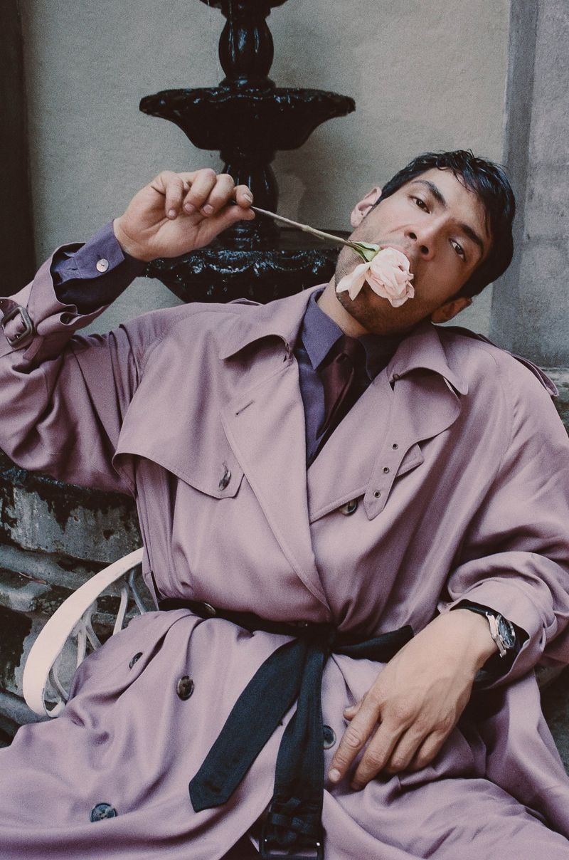 Actor Diego Calva in a pale lavender trench coat holding a pale pink rose in his mouth for a Man About Town editorial by Jacques Burga.
