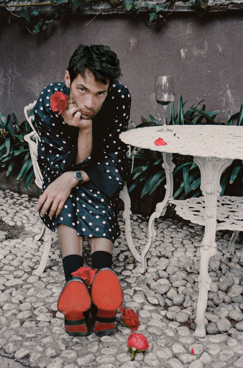 Actor Diego Calva in a polka-dot robe holding a rose while seated outdoors near a white table for Man About Town.
