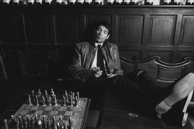 Diego Calva seated in a dark wood interior with feet up, wearing a leather jacket and tie, beside a chessboard.