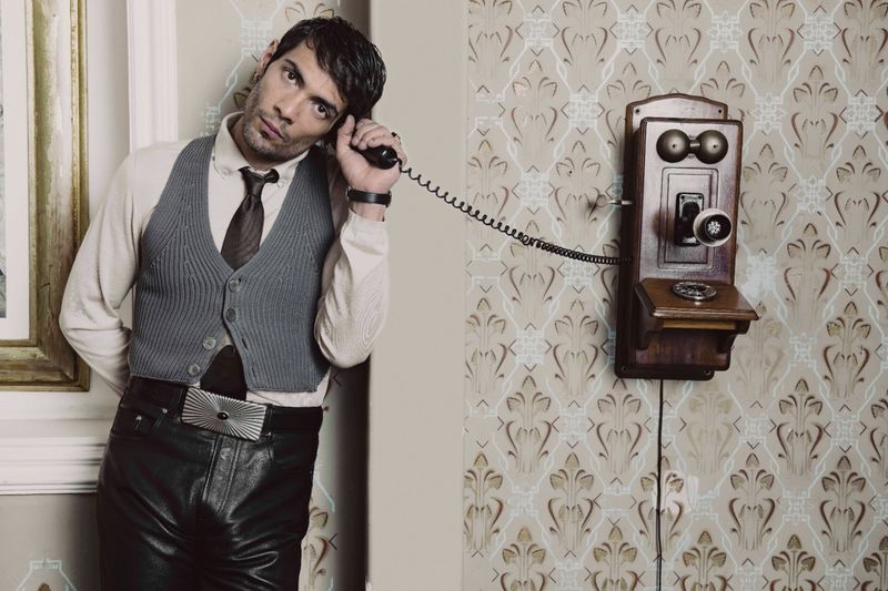 Diego Calva wearing a grey vest and black leather trousers holding an antique wall-mounted telephone receiver