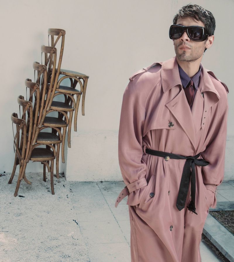 Diego Calva wearing a pink trench coat and sunglasses with a stack of wooden chairs in the background