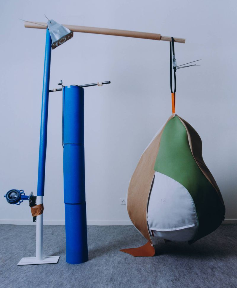 Hanging leather sculpture in green, white, and tan suspended from a makeshift wooden structure against a neutral wall.