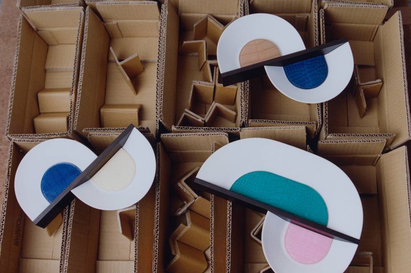 Overhead view of sculptural white objects with colored leather inserts arranged in open cardboard shipping boxes