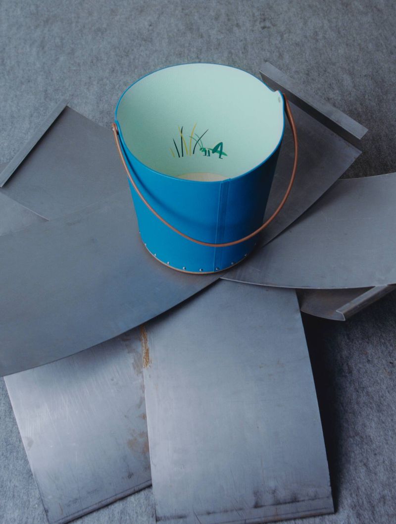 Blue bucket with leather handle resting on metallic sheets arranged radially on a grey textured surface