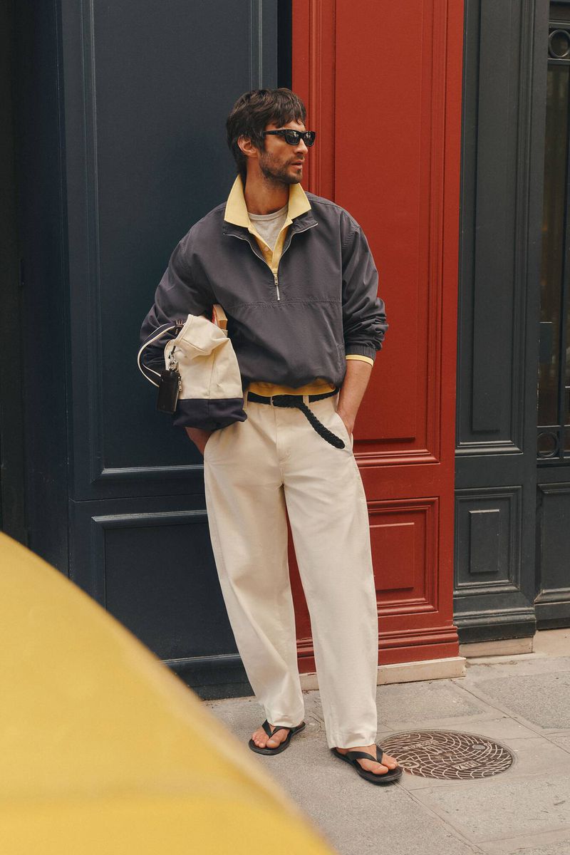 Man wearing sunglasses, grey jacket over yellow polo, and white trousers holding a canvas bag in front of a colorful storefront.