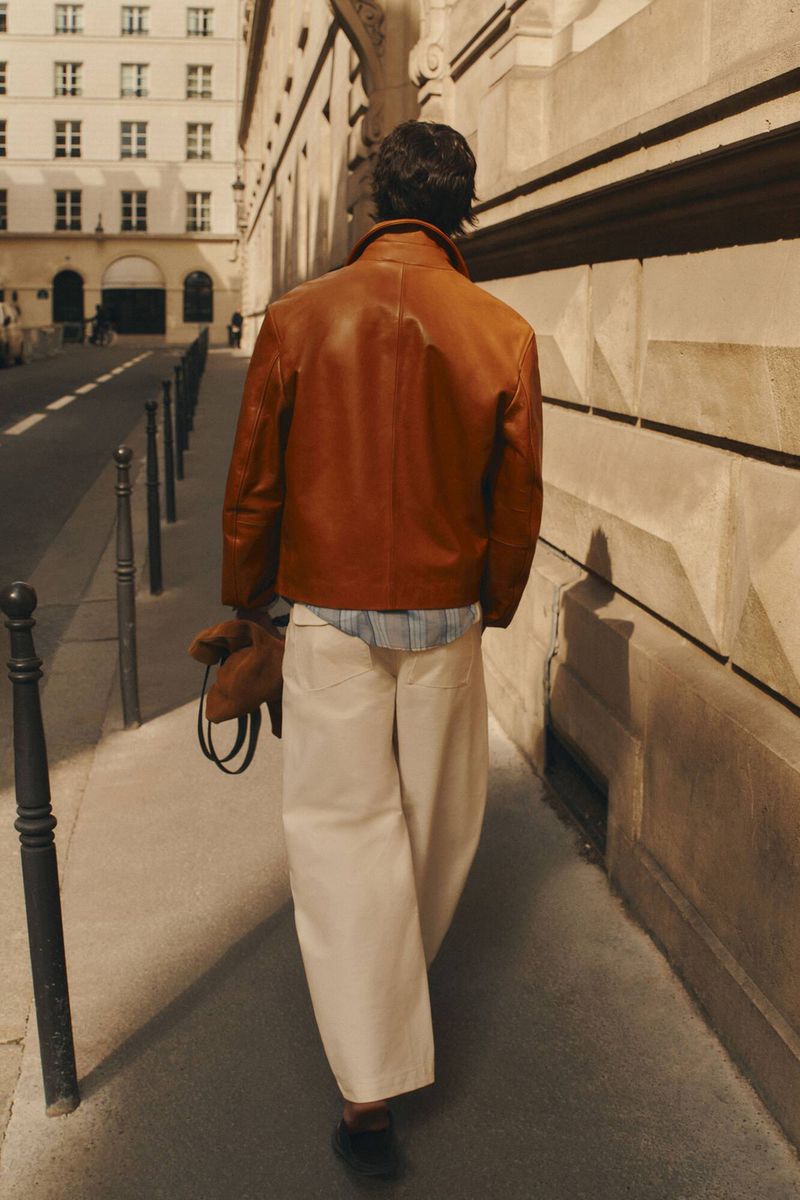 Man wearing a brown leather jacket and cream trousers walking away down a city sidewalk