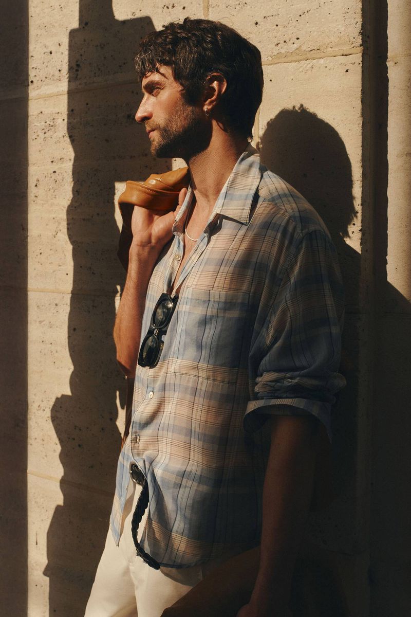 Portrait of a stylish adult male with a beard wearing a plaid shirt and sunglasses, standing outside with a tan jacket over his shoulder.