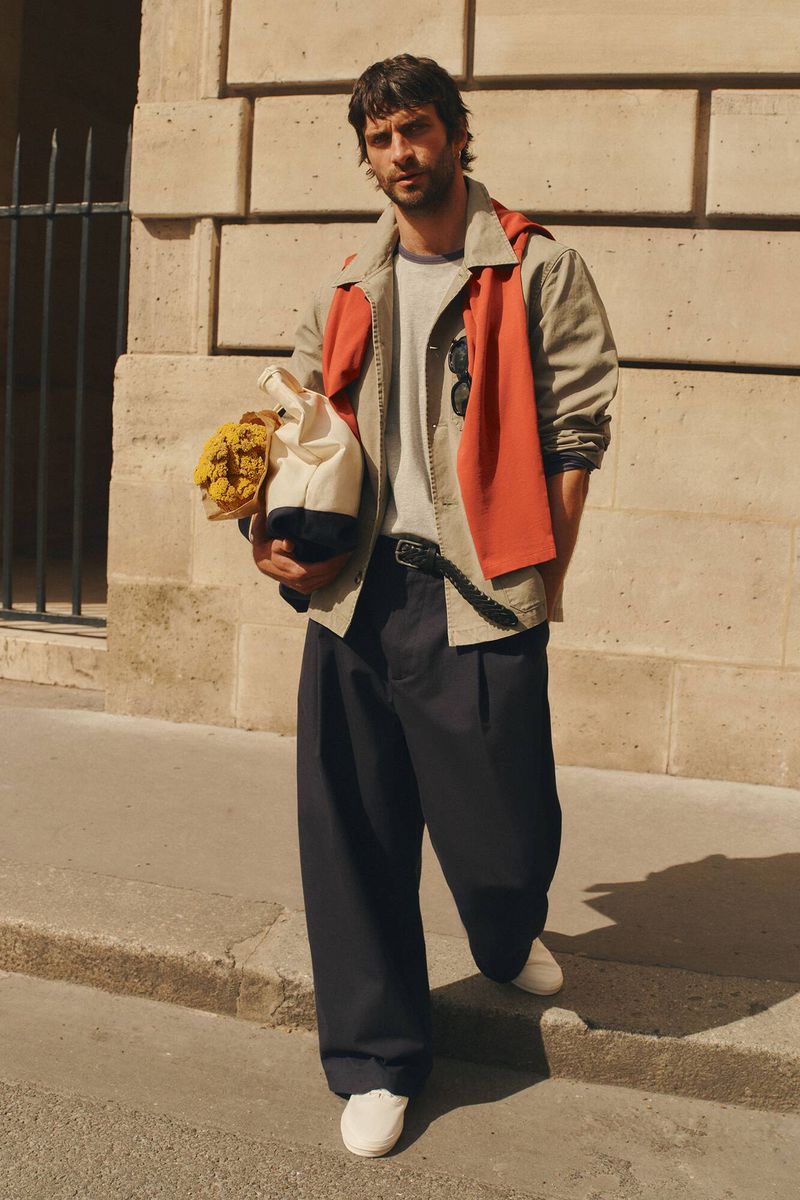 Man wearing a beige jacket and dark trousers holding a canvas bag with flowers standing outdoors on a sunny street