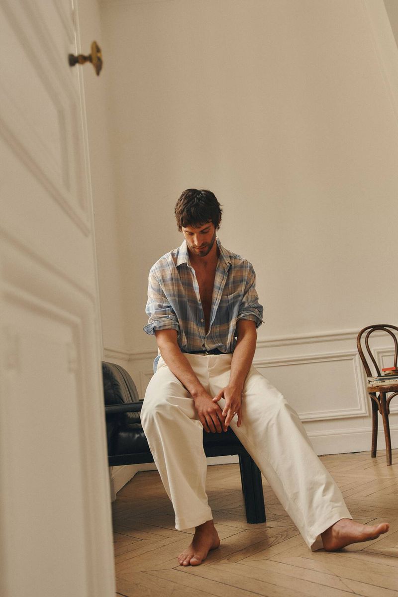 A man wearing an open plaid shirt and light trousers sitting barefoot on a chair in a room with wooden floors.