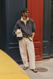 Man wearing sunglasses, grey jacket over yellow polo, and white trousers holding a canvas bag in front of a colorful storefront.