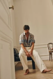 A man wearing an open plaid shirt and light trousers sitting barefoot on a chair in a room with wooden floors.