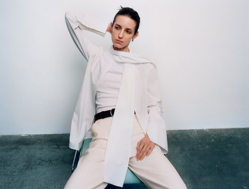 Model seated wearing a layered white shirt and light trousers against a neutral background for Financial Times HTSI Magazine