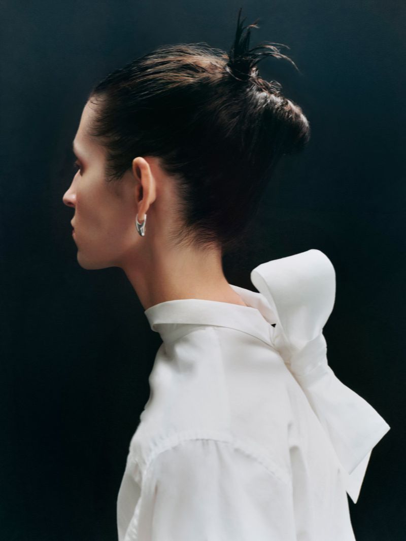 Profile portrait of a woman wearing a white shirt with a large bow on the back against a dark background.