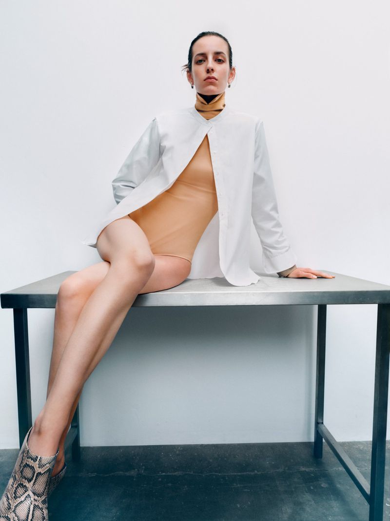 Indoor studio shot of a young adult woman in a bodysuit and white shirt, seated on a metal table, wearing snakeskin print shoes.