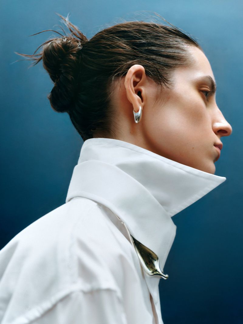 Profile portrait of a woman wearing a crisp white collared shirt and silver hoop earrings against a dark blue background.