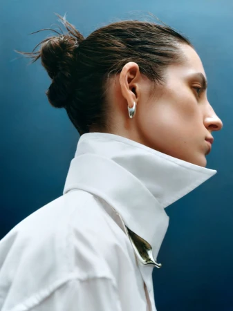 Profile portrait of a woman wearing a crisp white collared shirt and silver hoop earrings against a dark blue background.
