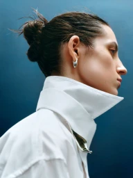 Profile portrait of a woman wearing a crisp white collared shirt and silver hoop earrings against a dark blue background.