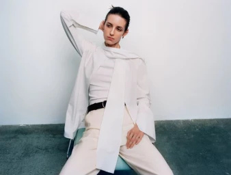 Model seated wearing a layered white shirt and light trousers against a neutral background for Financial Times HTSI Magazine