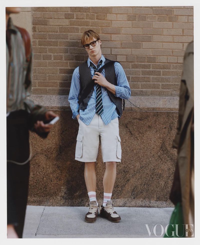 Man in preppy fashion style posing against brick wall, wearing glasses, striped shirt, vest, tie, and shorts with sneakers, outdoor setting.