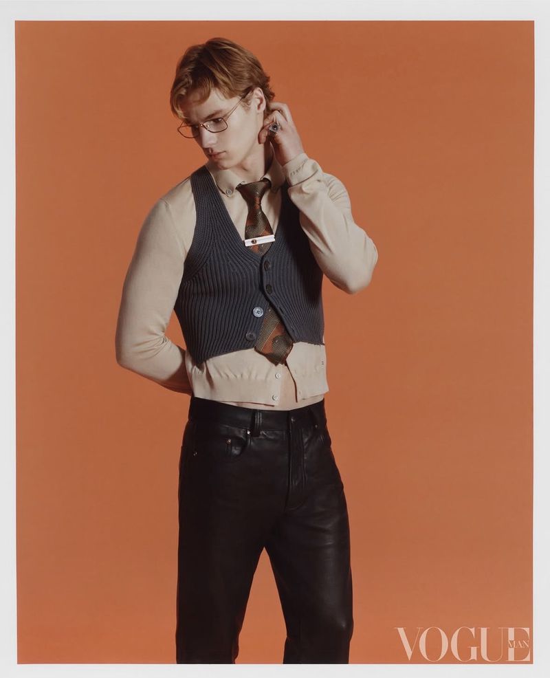 Young adult male fashion model poses against an orange backdrop wearing a shirt, vest, tie, glasses, and black pants in a studio.