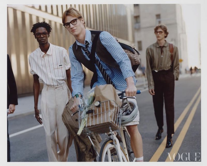 Four male models walking along a city street, with one riding a bicycle carrying a basket of flowers.