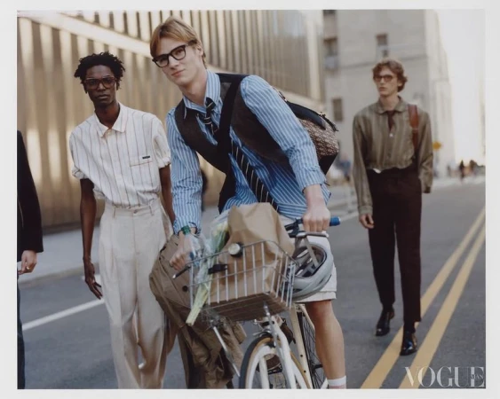 Four male models walking along a city street, with one riding a bicycle carrying a basket of flowers.
