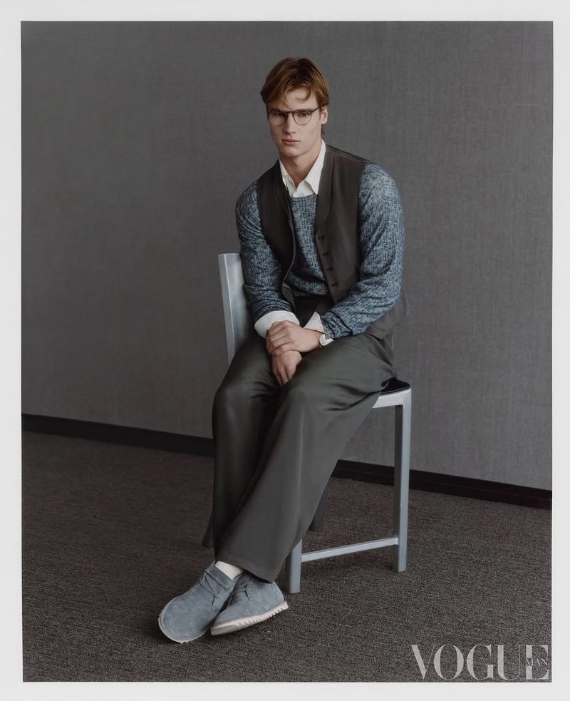 A male model wearing a grey vest, sweater, and trousers seated on a chair against a dark grey wall in a Vogue China Men editorial.