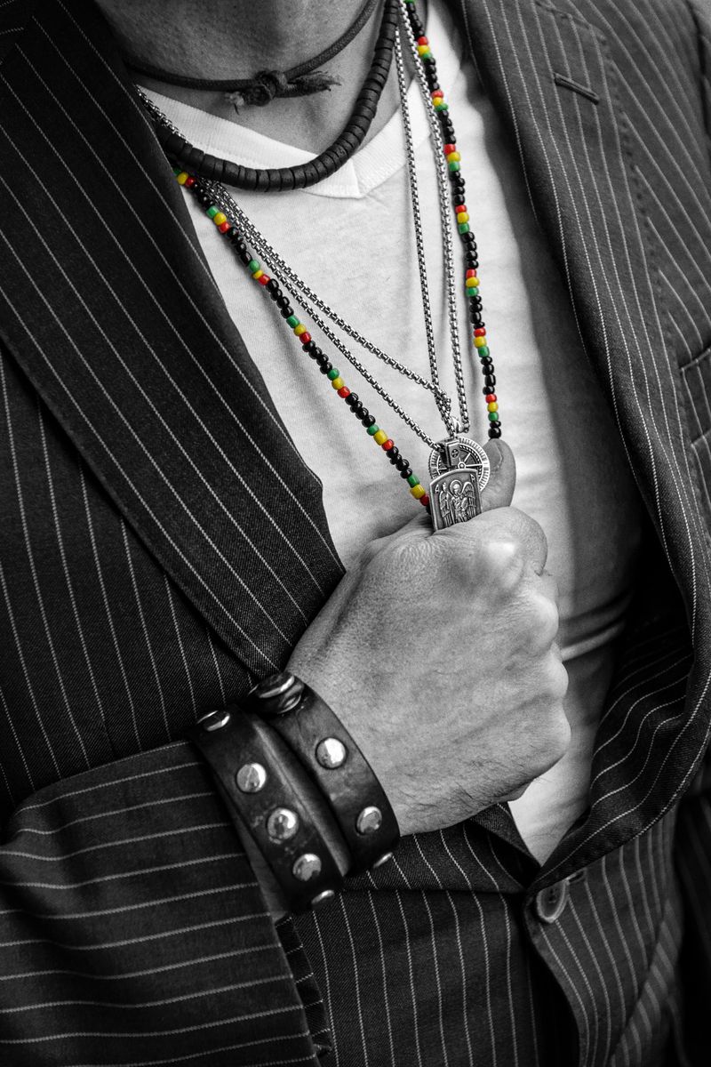 Man wearing a pinstripe blazer, beaded necklaces, and a leather bracelet strikes a casual pose in a monochromatic portrait.