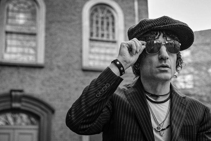Black and white portrait of a man in a pinstripe suit, newsboy cap, and sunglasses, looking off-camera with a brick building backdrop.