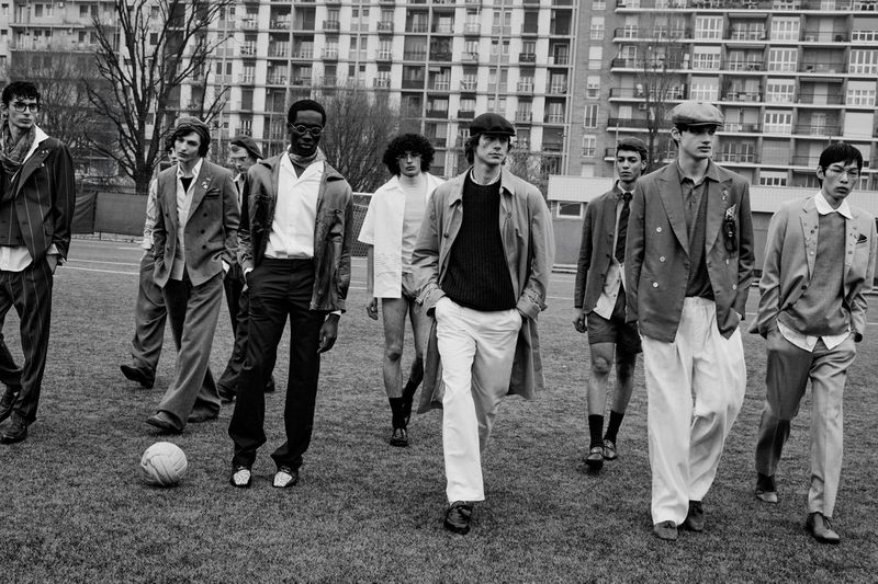 Group of male models in tailored menswear walking across a sports field with a soccer ball, set against apartment buildings.
