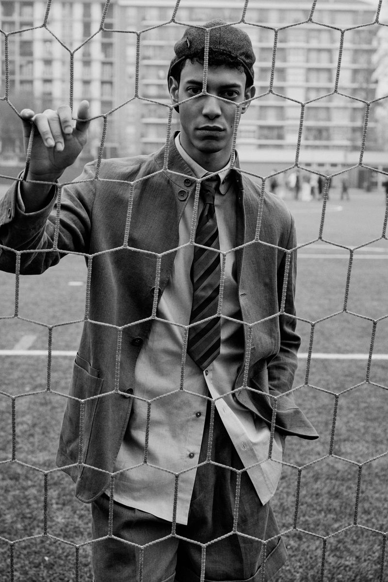 Young man in a suit jacket and cap posing behind a soccer goal net on a field for L'Officiel Hommes Italia.
