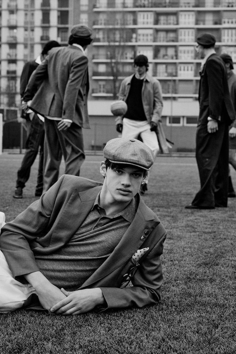 Model in flat cap and suit jacket reclining on a sports field with other figures standing in background