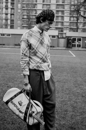 Monochrome fashion editorial image of a male model wearing a plaid cap and sweater, holding a sports bag on a field
