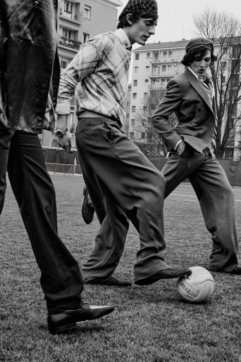 Black and white editorial image of multiple men playing soccer in vintage attire outdoors near apartment buildings