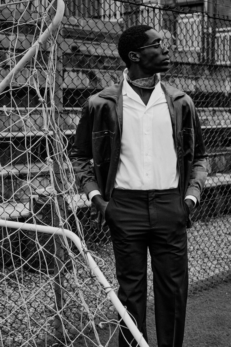 Black and white portrait of a man in a white shirt and dark jacket standing by a chain-link fence and a soccer goal