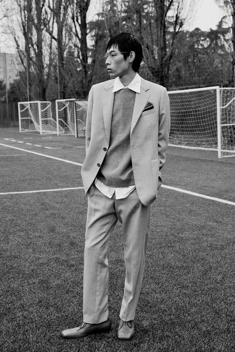 Asian male model in light grey suit standing on grass soccer field with goalposts and trees, black and white fashion editorial.