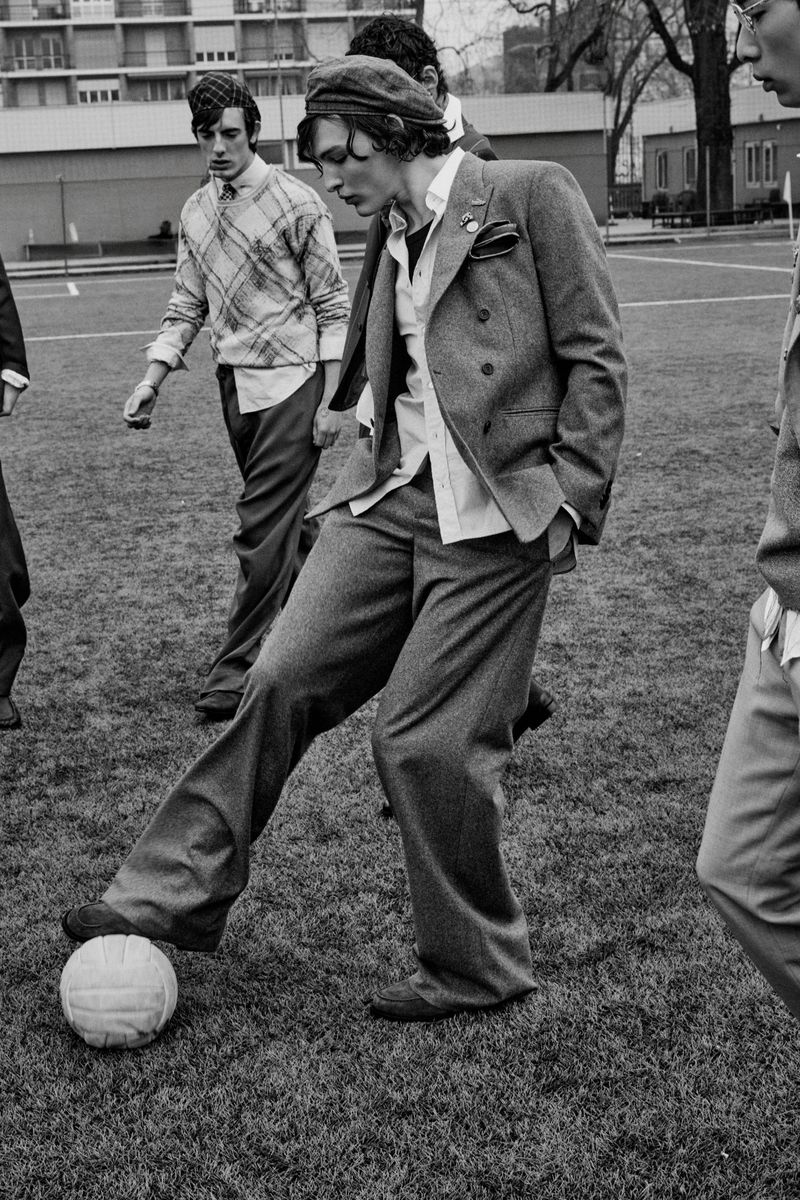 Monochrome fashion editorial image of several men in tailored suits and caps playing soccer on a field