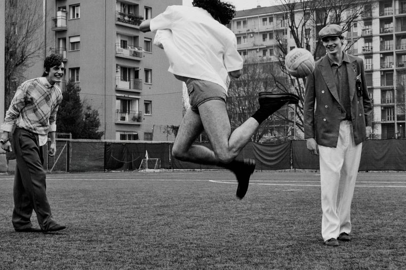 Black and white editorial image of a young man jumping to kick a soccer ball, watched by two other men on a pitch.